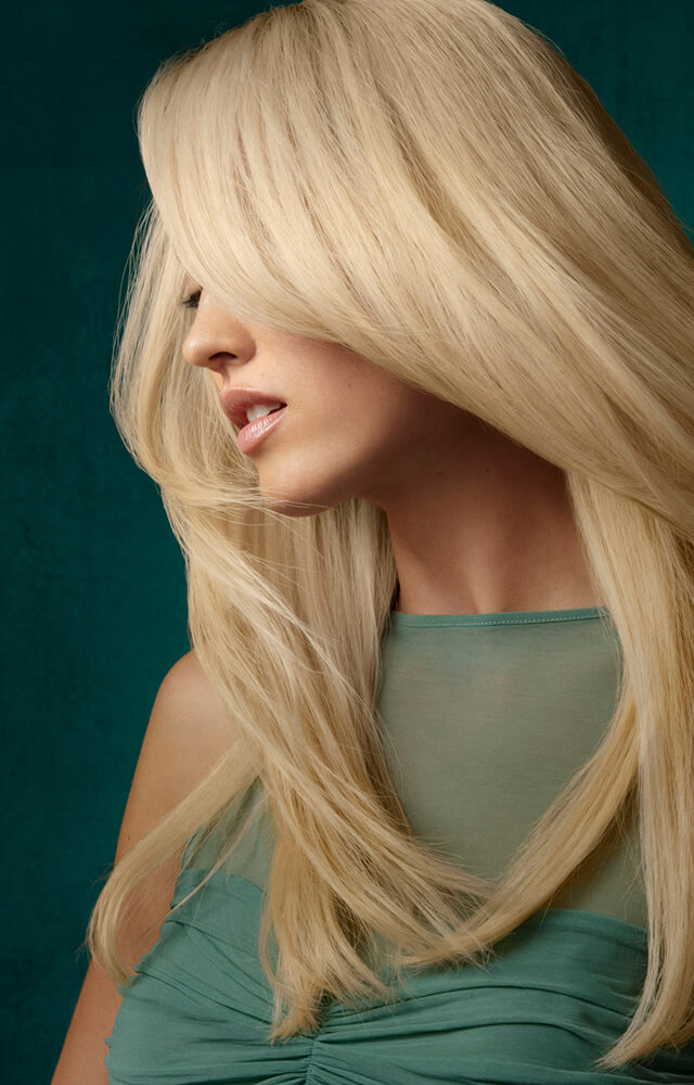 A woman with long, straight platinum blonde hair color looks to the side. She is wearing a sleeveless teal top against a dark green background.
